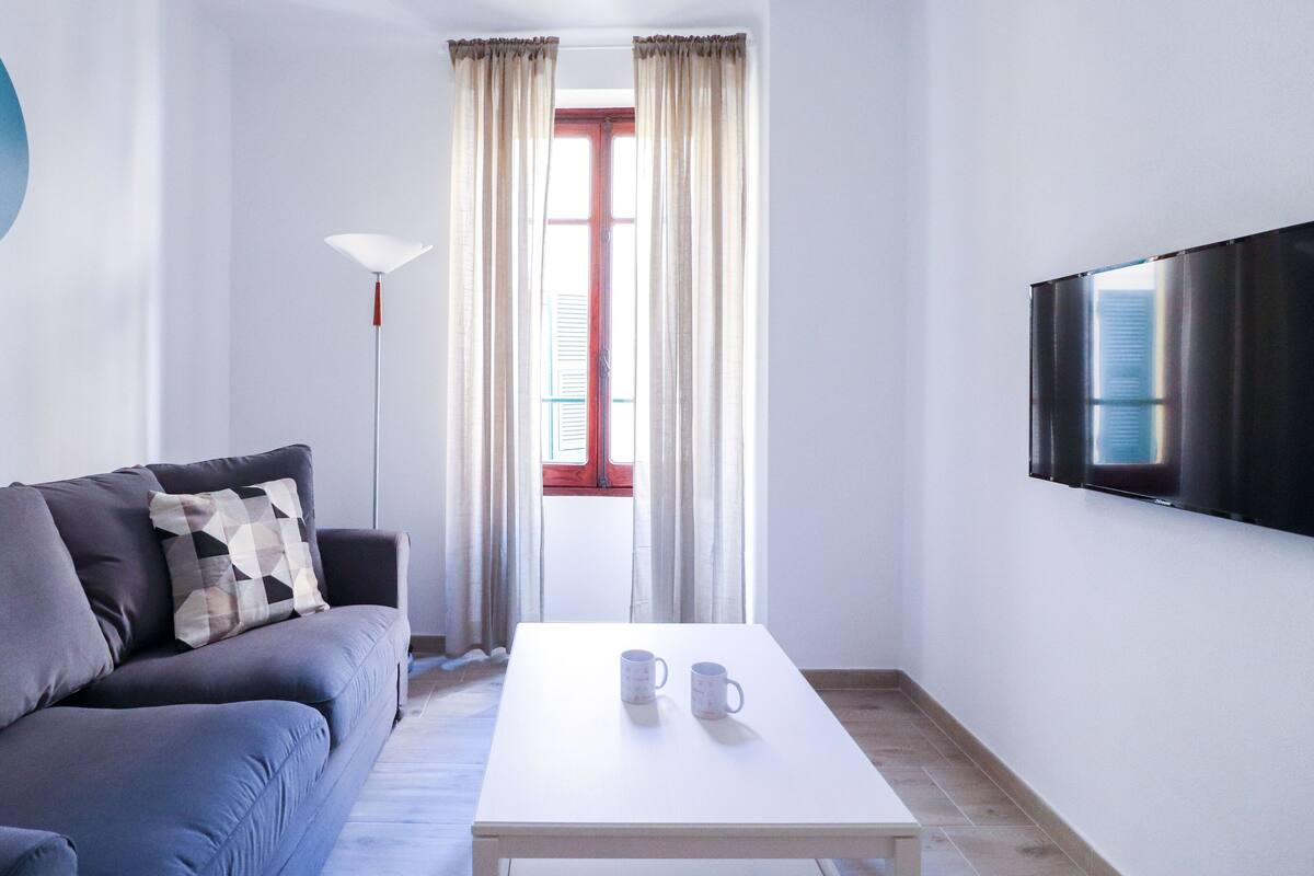 Un salon moderne avec un canapé gris, une table basse blanche sur laquelle reposent deux tasses, un lampadaire et un téléviseur fixé au mur.