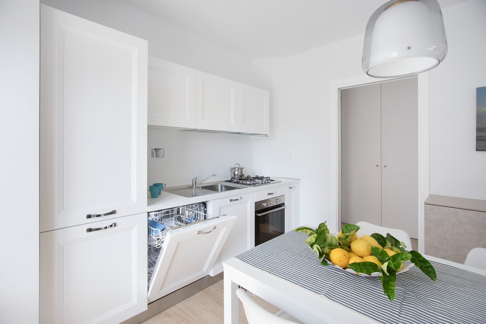 A modern kitchen with white cabinetry, a built-in dishwasher, a stove, and a sink. There is a dining table with a striped tablecloth and a bowl of lemons.