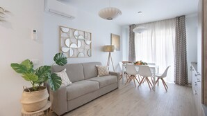 Una sala moderna con sofá, mesa de comedor y sillas. Hay una planta grande, un adorno de pared y una lámpara de pie.