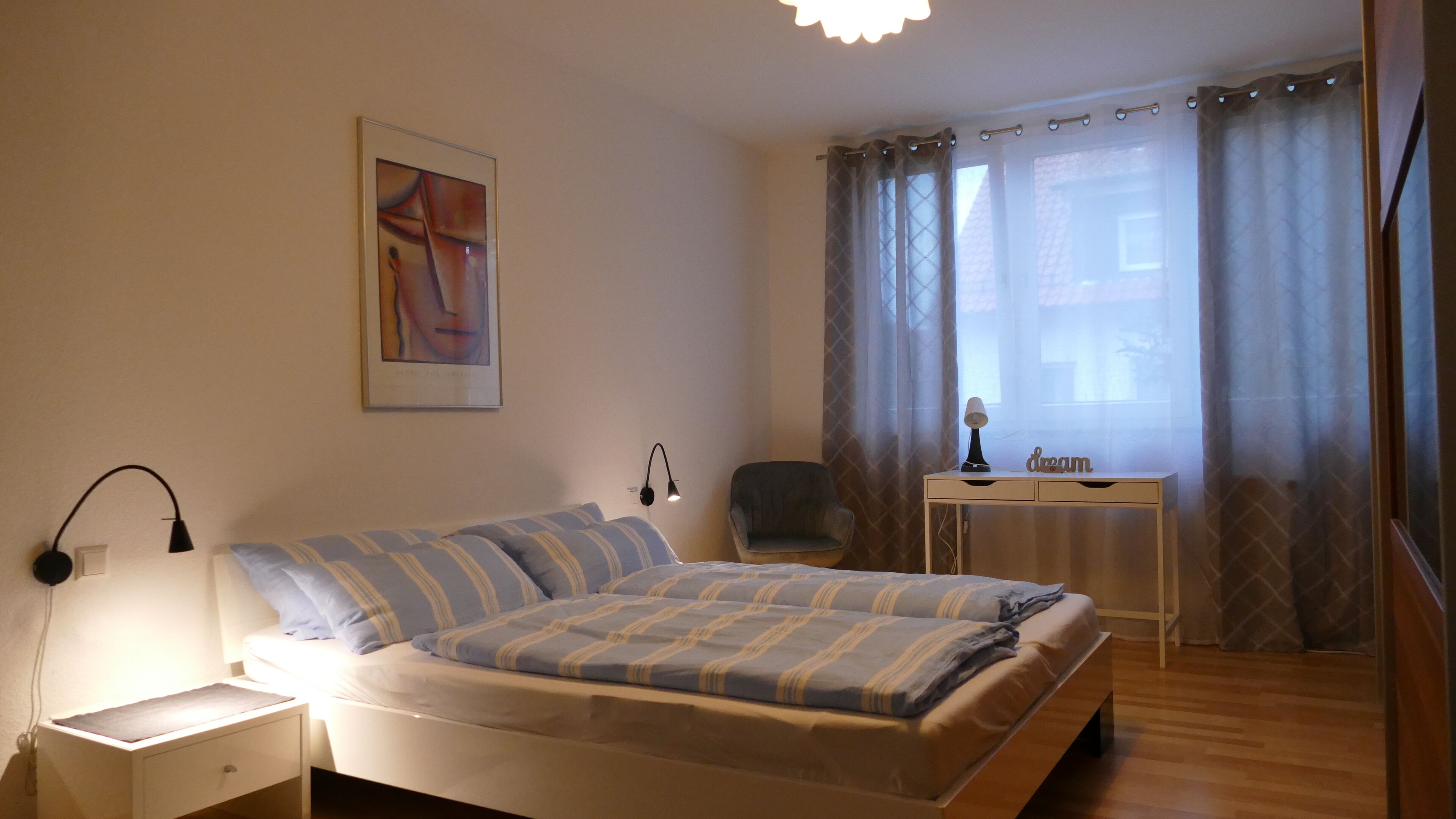 Une chambre à coucher comprenant un lit, une table de chevet, une lampe, une chaise, un bureau et des rideaux.
