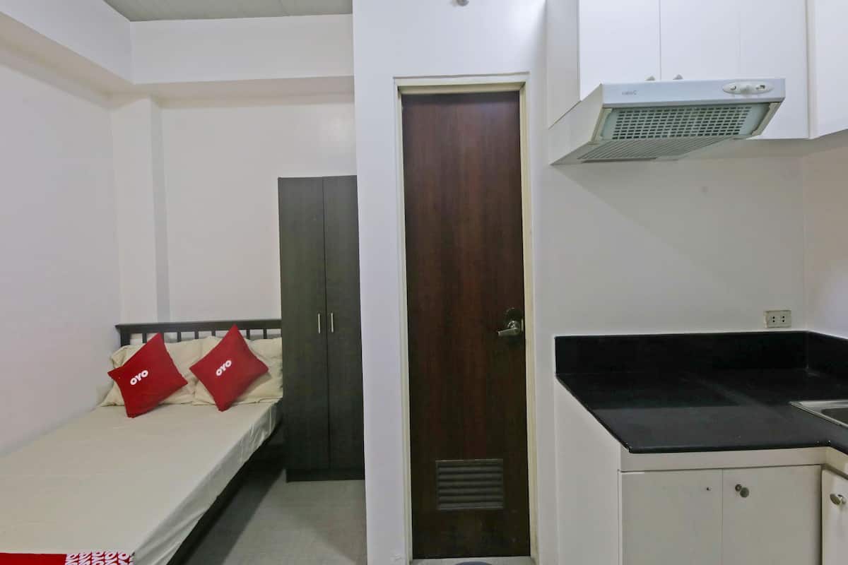 A small, compact room with a single bed, a wardrobe, a kitchenette, and a door leading to another room.