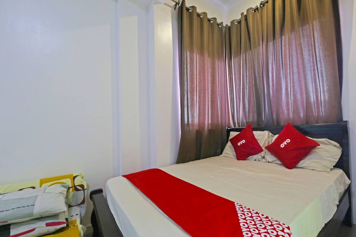 A hotel room with a bed, two red pillows, a red throw, and a yellow trolley with towels.