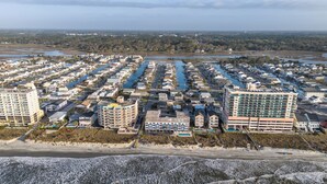 An aerial view of a coastal residential area with multiple buildings, including a prominent hotel with a large balcony and a view of the beach.