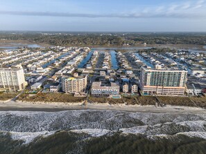 Ett flygbildsbild av ett bostadsområde vid kusten med flera byggnader, inklusive ett framstående hotell med en stor balkong och utsikt över stranden.