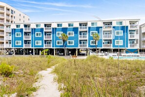 Un hotel frente a la playa con una hilera de edificios azules y blancos, un camino de arena que lleva a la entrada y un cielo despejado.