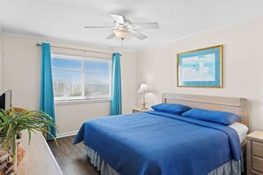 Ein Schlafzimmer mit Bett, Deckenventilator, Fenster mit Vorhängen, Nachttischlampe und einem gerahmten Bild an der Wand.