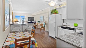 A compact kitchen with white cabinets, a stove, and a refrigerator. There is a dining area with a table and chairs. - Oceanfront 2BR • Balcony Views  & Oceanside Pool • Dog Friendly • A Summer Place B8 (North Myrtle Beach)