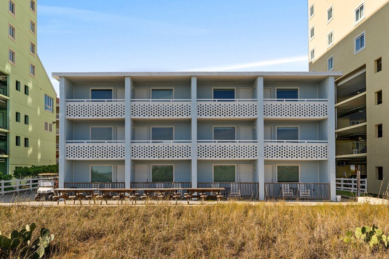 A multi-story building with balconies, a flat roof, and a parking area with benches.