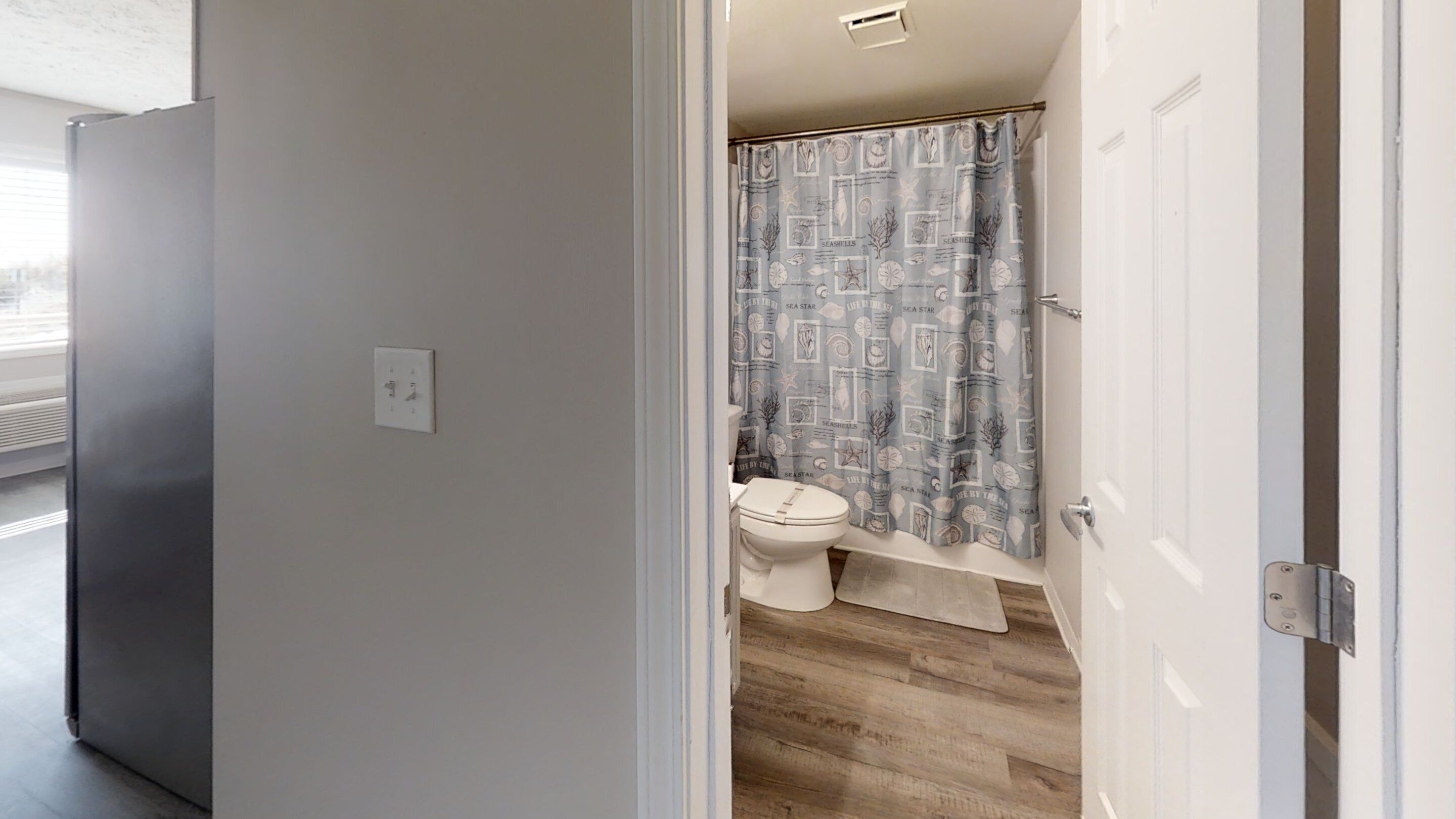 A bathroom with a toilet, a shower curtain with a sea-themed pattern, and a wooden floor.