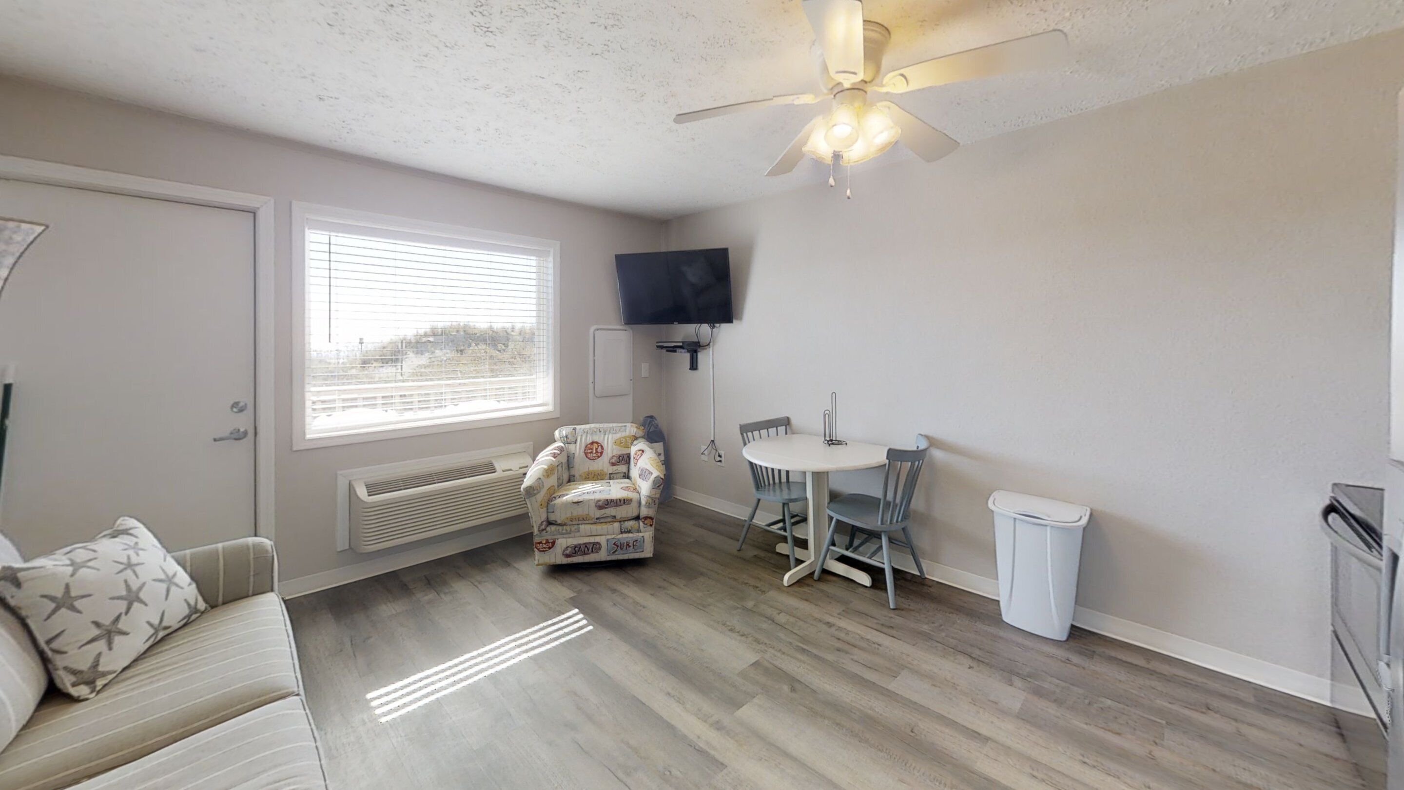 A compact living space with a sofa, a patterned armchair, a dining table with chairs, a ceiling fan, a TV mounted on the wall, and a window with blinds.