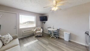 Un espacio habitable compacto con sofá, un sillón estampado, una mesa de comedor con sillas, un ventilador de techo, un televisor montado en la pared y una ventana con persianas.