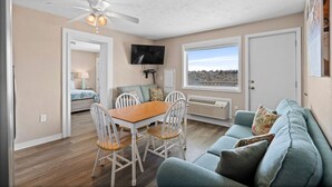 A spacious living area with a dining table, chairs, a sofa, a ceiling fan, a TV, and a large window with a view.