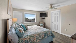 A bedroom with a bed, a TV mounted on the wall, a ceiling fan, a window with a view, and a door.