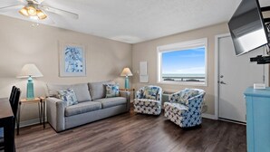 A living room with a grey sofa, two patterned armchairs, a ceiling fan, a TV, and a window with a view of the ocean. - Pet-Friendly Oceanfront 2-BR on Top/3rd Floor – Stunning Cherry Grove Beach Views - Blue Mist 302 (North Myrtle Beach)
