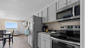 A compact kitchen with a refrigerator, microwave, stove, and sink. There is a dining area with a table and chairs. - Pet-Friendly Oceanfront 2-BR on Top/3rd Floor – Stunning Cherry Grove Beach Views - Blue Mist 302 (North Myrtle Beach)