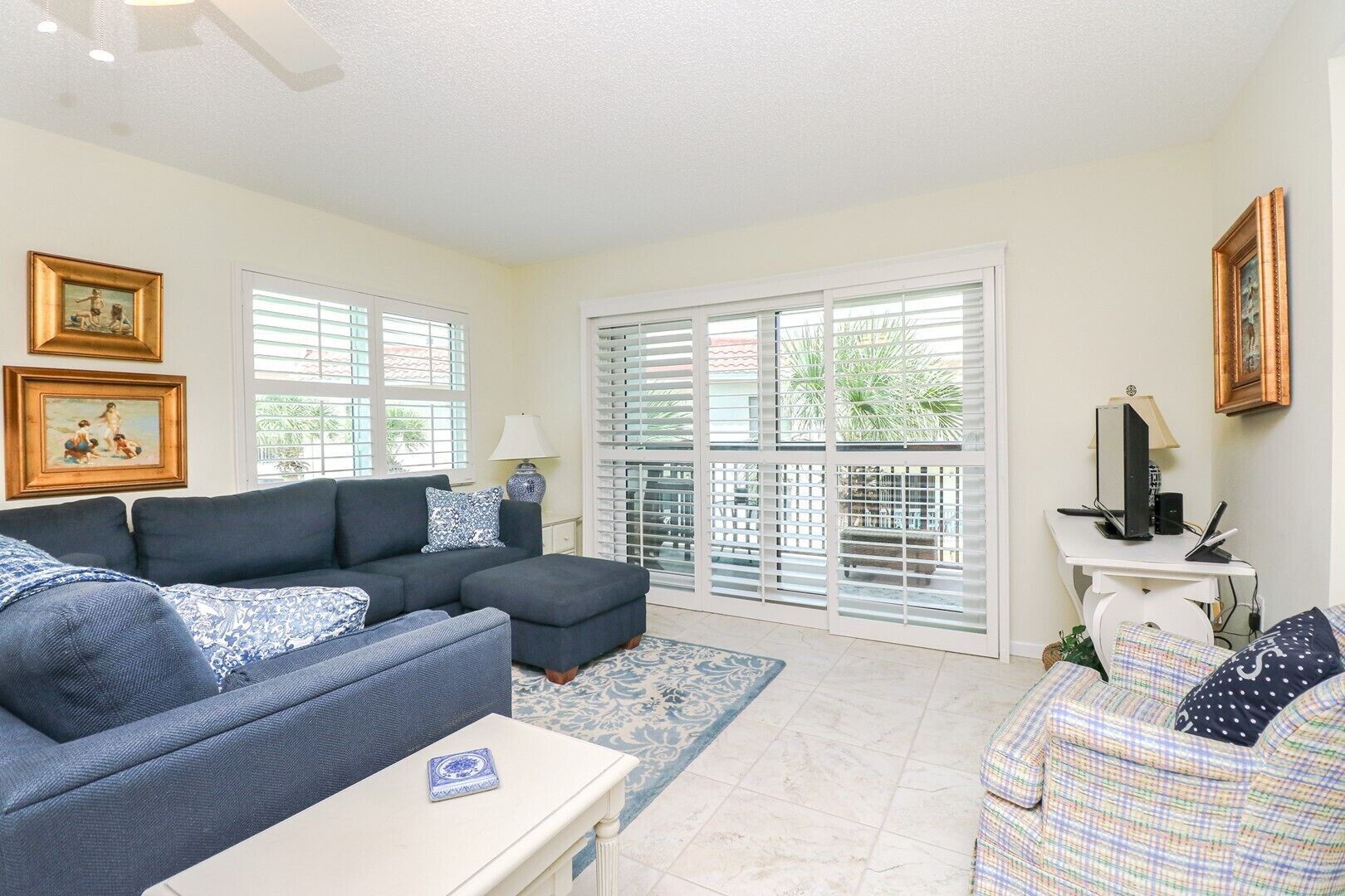 A living room with a sectional sofa, a small table, and a desk with a computer. There are windows with blinds and a door with glass panels.