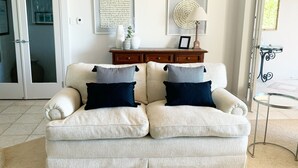 Un sofá beige con dos almohadones oscuros, una mesita de madera con una lámpara y una mesa de vidrio con estructura metálica.