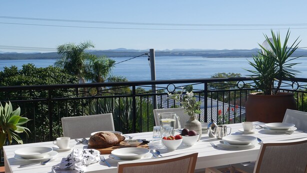 Una mesa de comedor en un balcón con vista al mar y a las montañas.