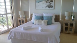 Una cama bien hecha con sábanas blancas y dos toallas enrolladas, flanqueada por dos mesitas de noche con lámparas y libros, en una habitación con un ventanal grande y un cuadro con motivos de playa.