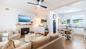Ein Wohnzimmer mit Kamin, ein Essbereich mit Tisch und Stühlen, eine Küche mit Kühlschrank und Spüle sowie eine große Uhr an der Wand.