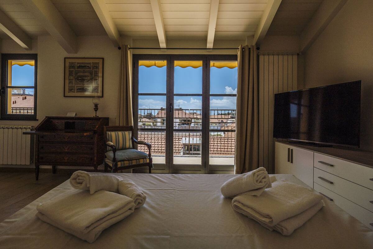 A hotel room with a bed, a TV, a desk, a chair, and a view of the city through large windows.