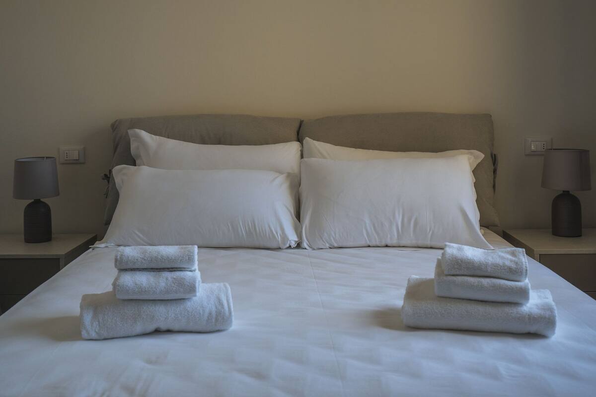A neatly made bed with white linens and pillows, two bedside tables with lamps, and folded towels on the bed.