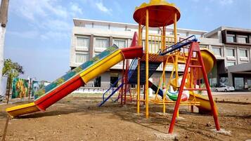 A playground with slides and climbing structures, set against a building with multiple windows and balconies.