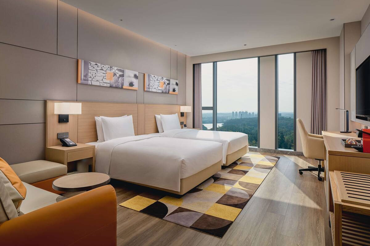 A modern hotel room with a large bed, two bedside tables, a desk, and a chair. There are large windows offering a city view.