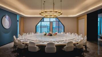 A modern conference room with a large oval table, white chairs, a chandelier, and a city view through large windows.