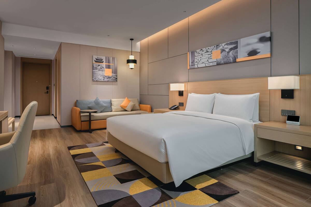 A modern hotel room with a large bed, a sofa, a desk, and a chair. There are two bedside tables with lamps, a wall-mounted TV, and artwork on the walls.