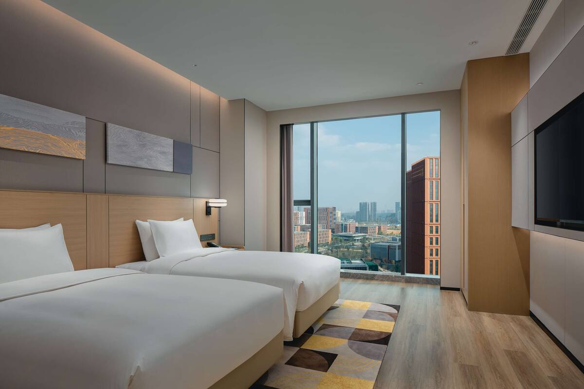 A hotel room with two beds, a large window, a flat-screen TV, and a city view.