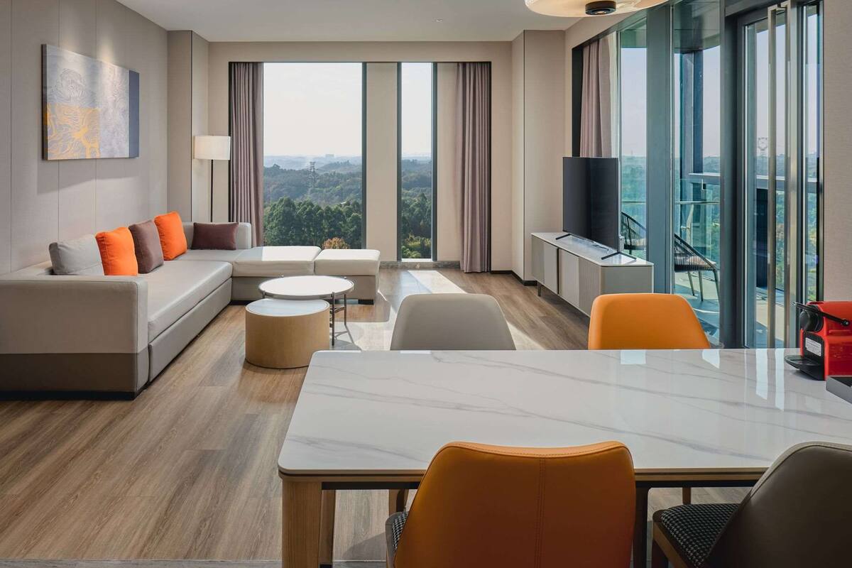 A modern hotel room with a sofa, ottoman, dining table, and chairs.