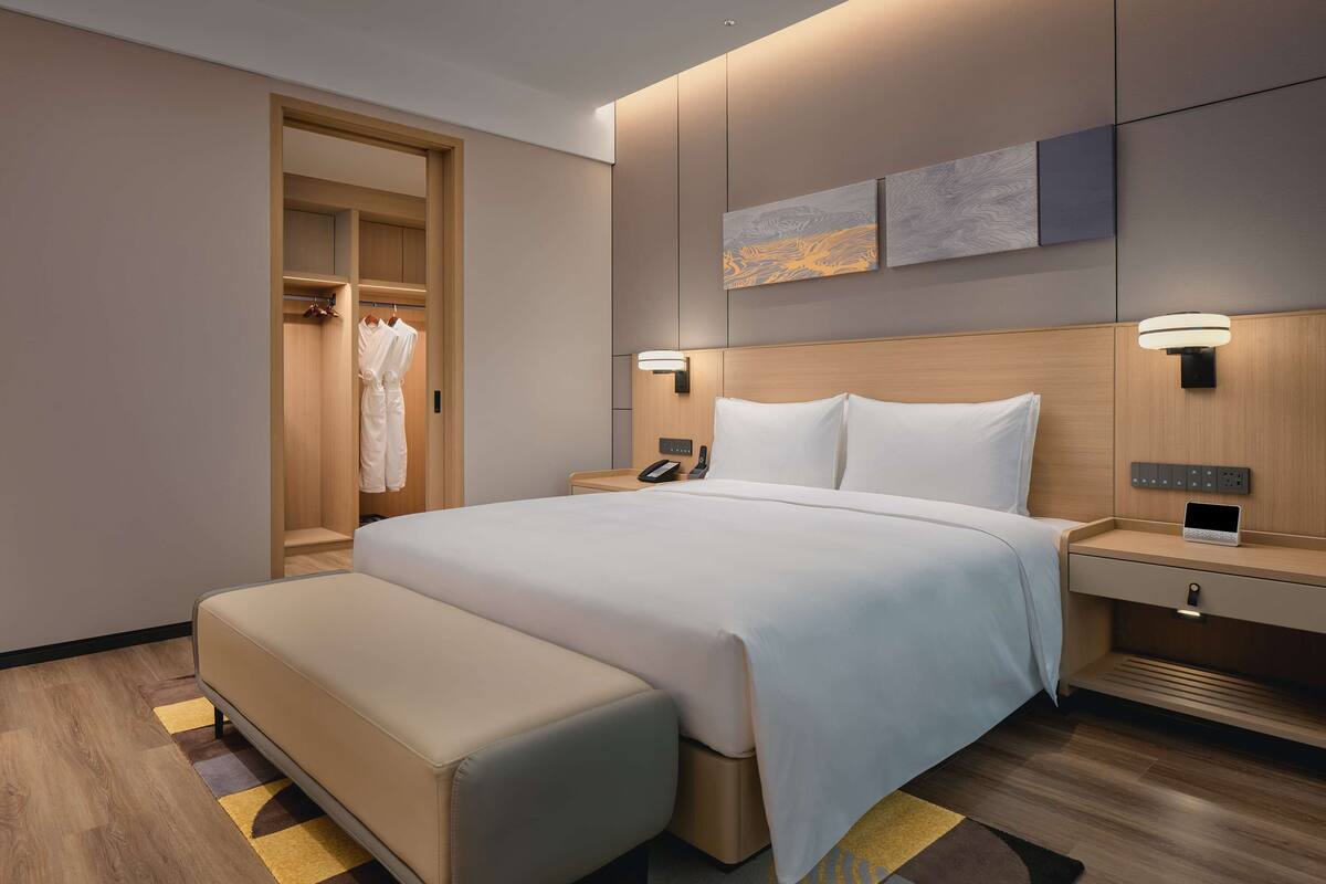 A modern hotel room with a large bed, a bedside table with a phone, a bedside lamp, a wooden headboard, a wardrobe, and a bench.