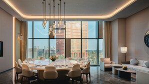 Un espace repas moderne avec une grande table, plusieurs chaises et un lustre. On aperçoit un canapé et une lampe au fond.