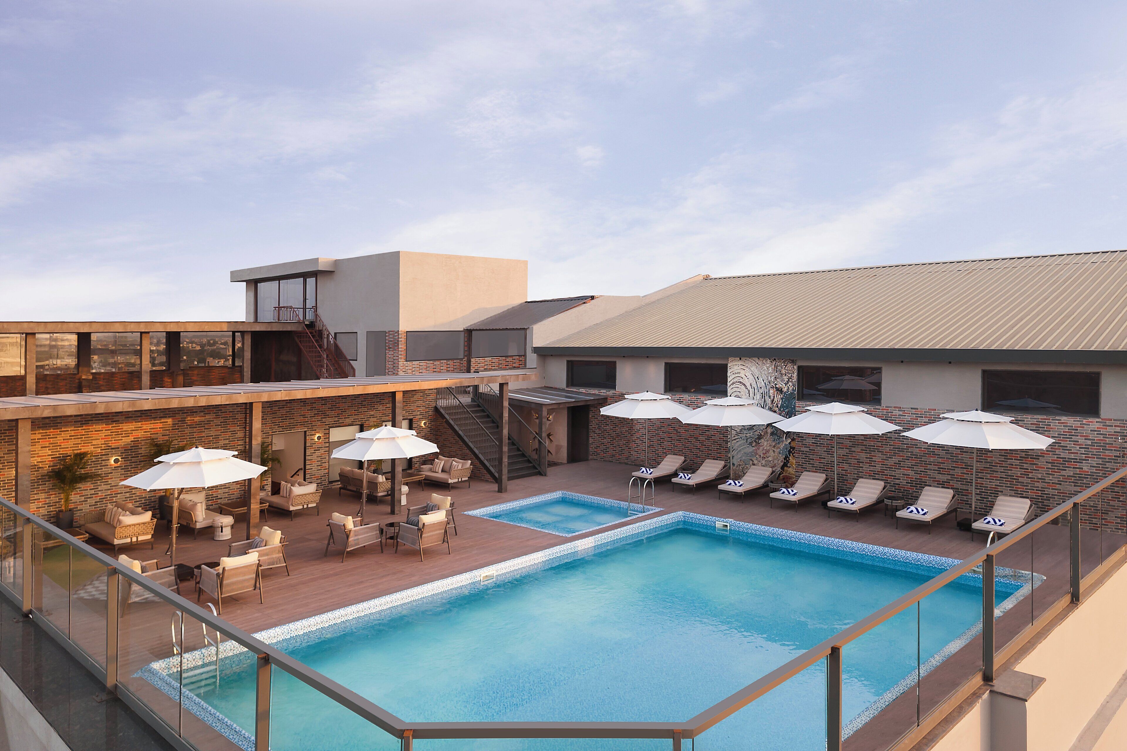 Una piscina en la azotea con sillones de descanso y sombrillas, una escalera y un edificio moderno con ventanas grandes.