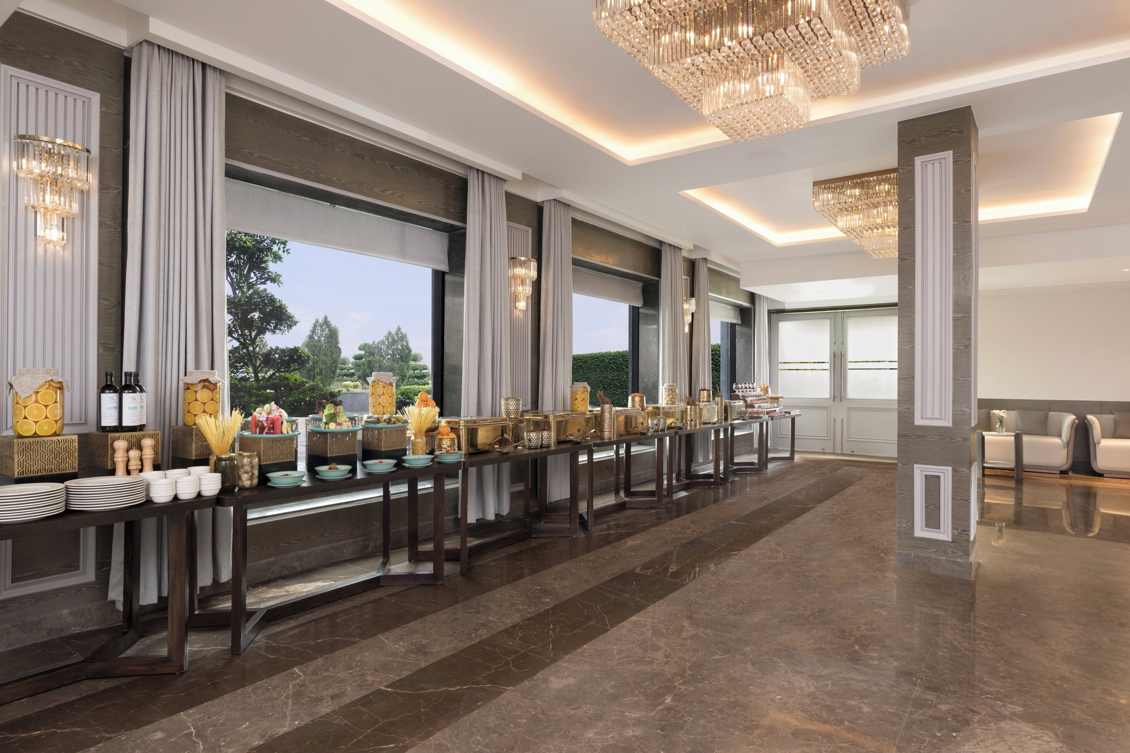 Una larga mesa de banquete con diversos platos y centros de mesa, ventanas amplias con cortinas y un candelabro.