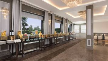 Una larga mesa de banquete con diversos platos y centros de mesa, ventanas amplias con cortinas y un candelabro.