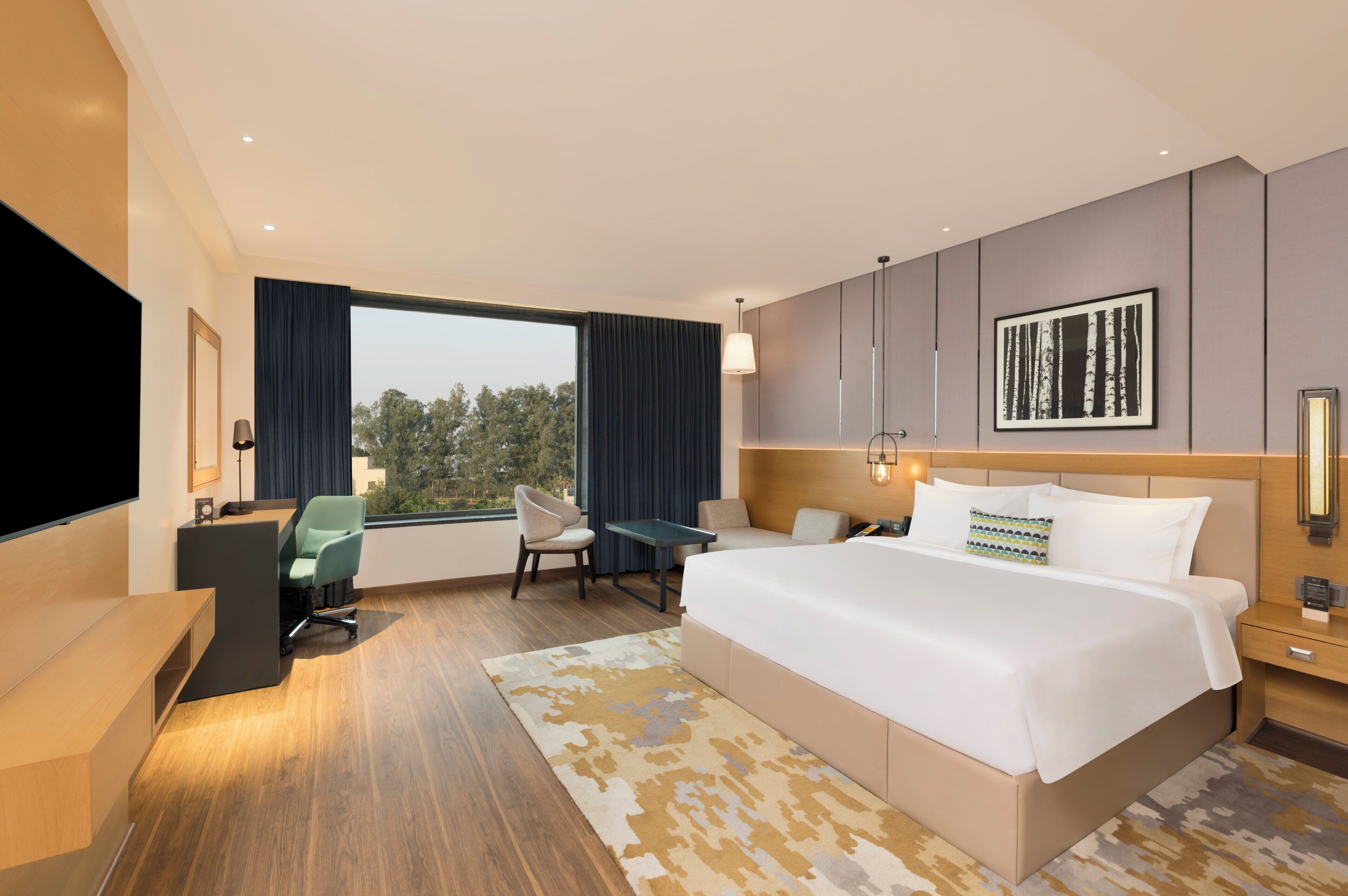 A modern hotel room with a large bed, a desk, a chair, a TV, and a large window.