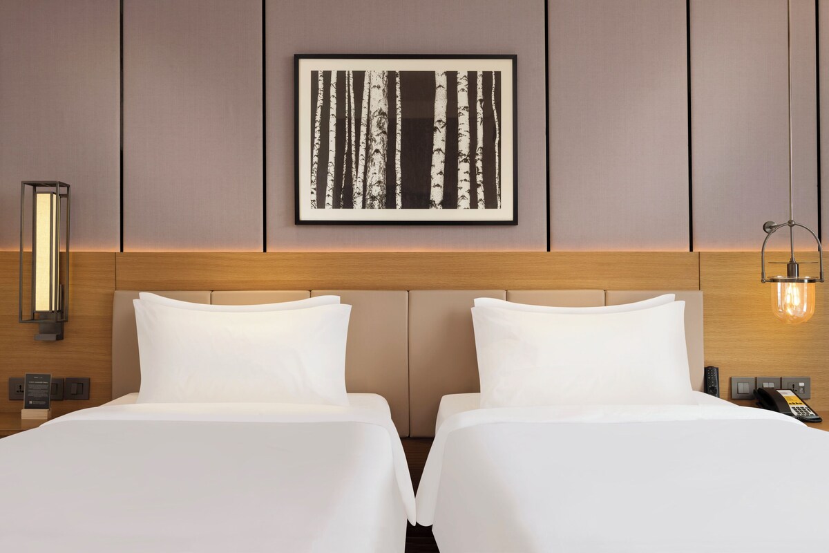 A hotel room with two beds, a framed picture on the wall, a bedside table with a lamp, and a phone on the nightstand.