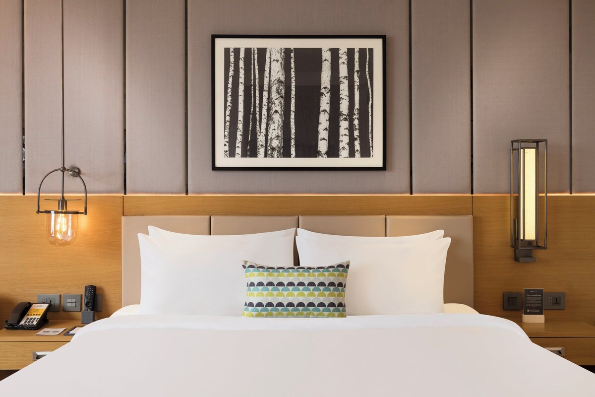 A hotel room with a bed, a framed picture on the wall, a bedside table with a lamp, and a phone.