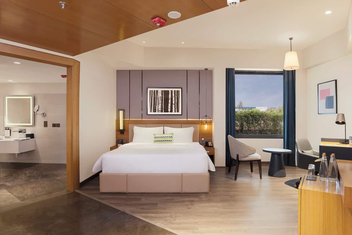 Ett modernt hotellrum med en säng, ett sängbord, en stol, ett skrivbord och ett stort fönster med utsikt.