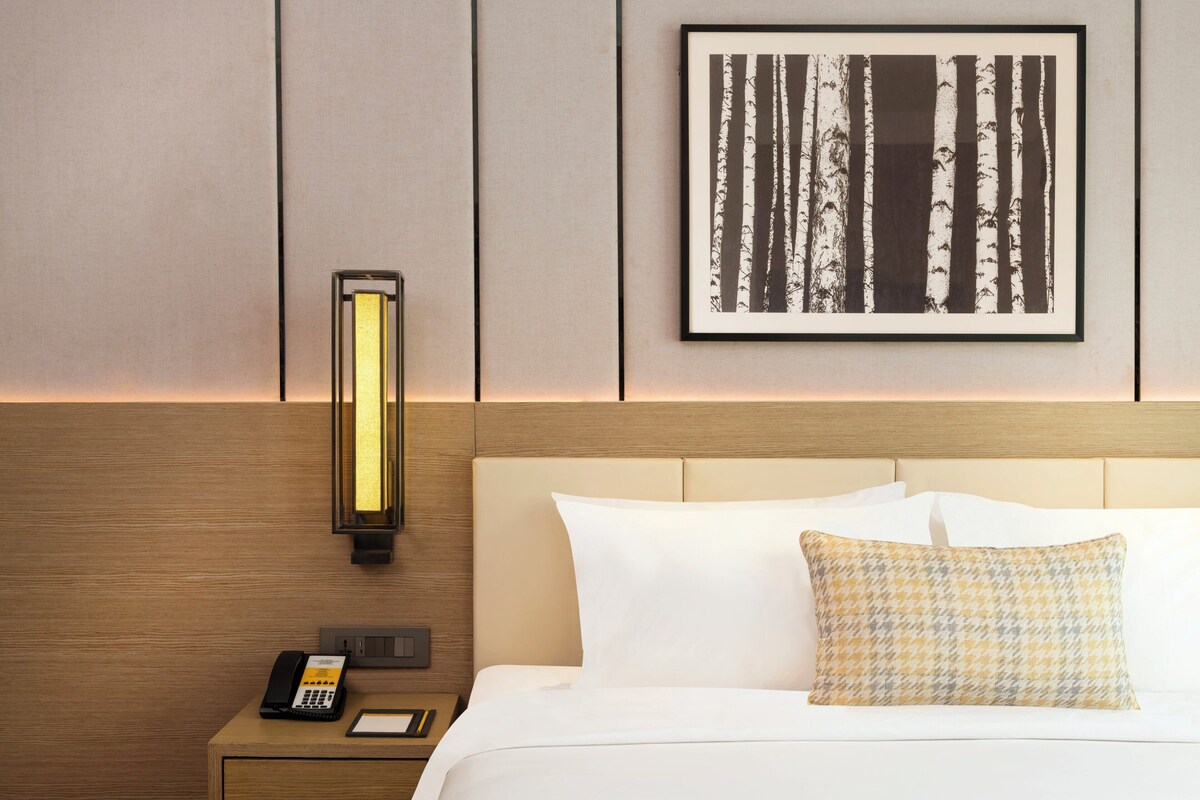 A hotel room with a bed, a nightstand with a phone, a wall-mounted light, and a framed picture.