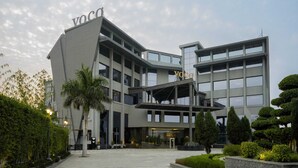 Un edificio moderno con un llamativo letrero 'Voco', que presenta una estructura de varios niveles con grandes ventanales y una zona de entrada ajardinada.