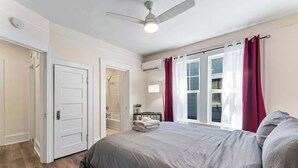 Un dormitorio con cama, ventilador de techo, ventana con cortinas, una puerta y una mesita de noche pequeña.
