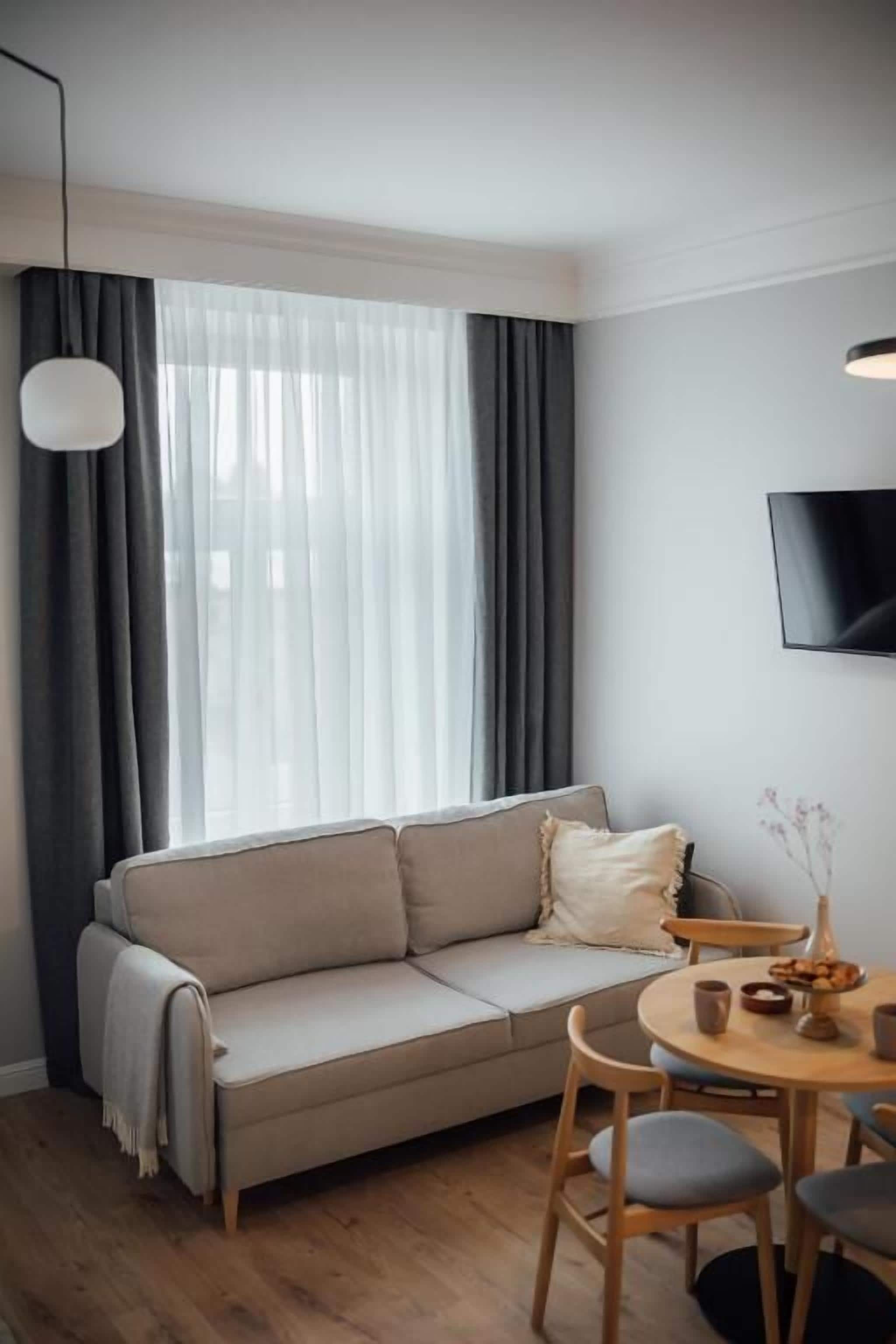 A modern living room with a sofa, a dining table with chairs, a flat-screen TV mounted on the wall, and a large window with sheer curtains.