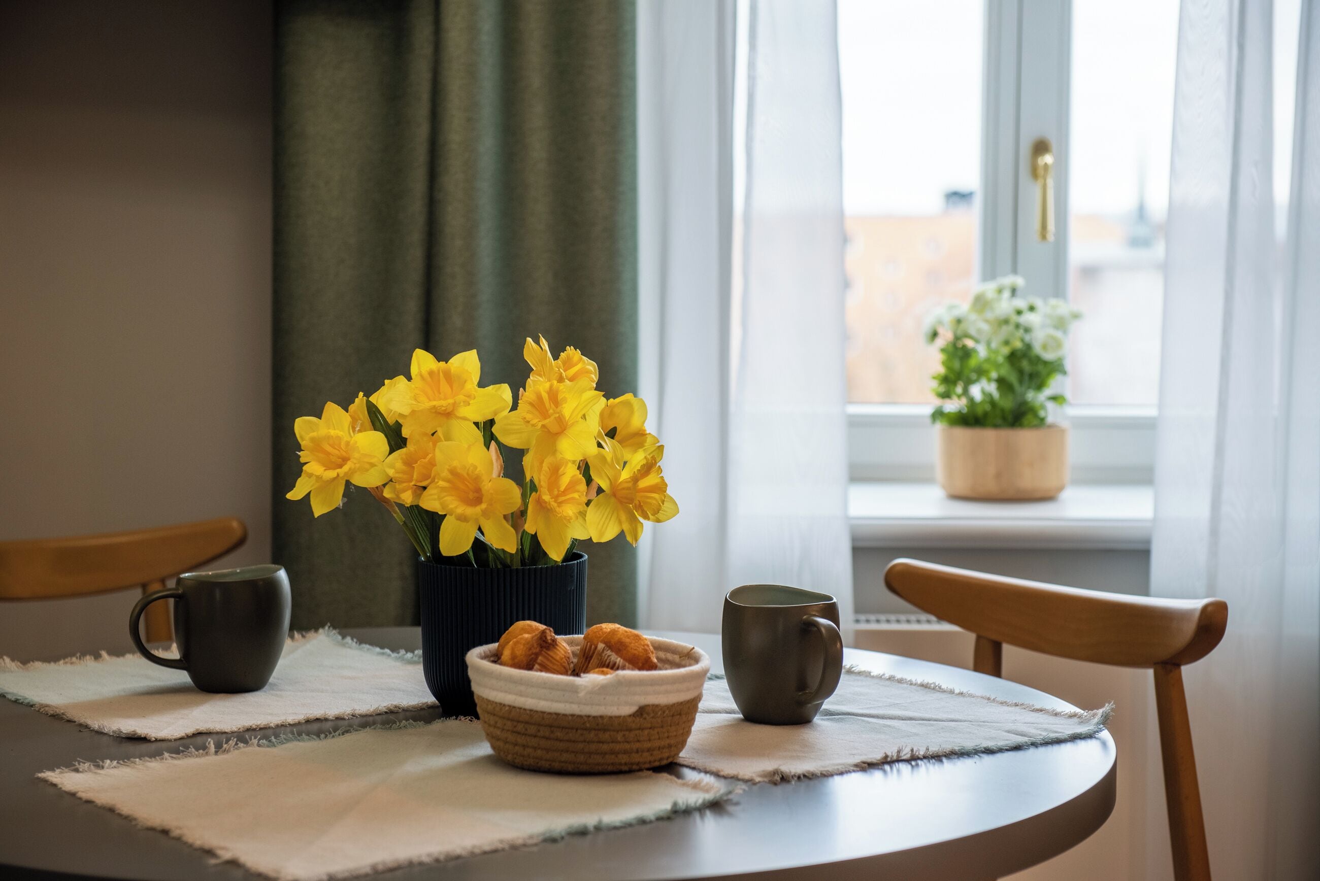 A table with a vase of yellow flowers, a bowl of pastries, and two mugs on a placemat.