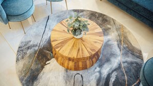 A round wooden coffee table with a plant on top, surrounded by a marble-patterned rug, in a room with blue upholstered chairs and a sofa.