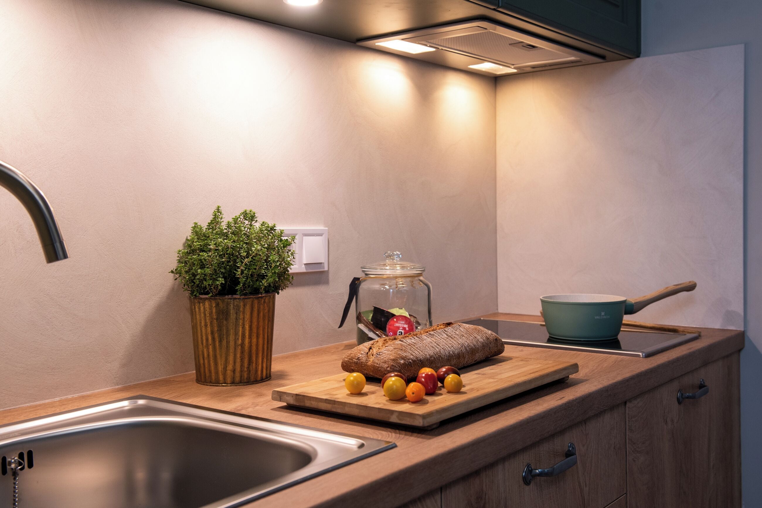 A kitchen with a wooden countertop, a sink, a potted plant, a loaf of bread, tomatoes, and a frying pan.