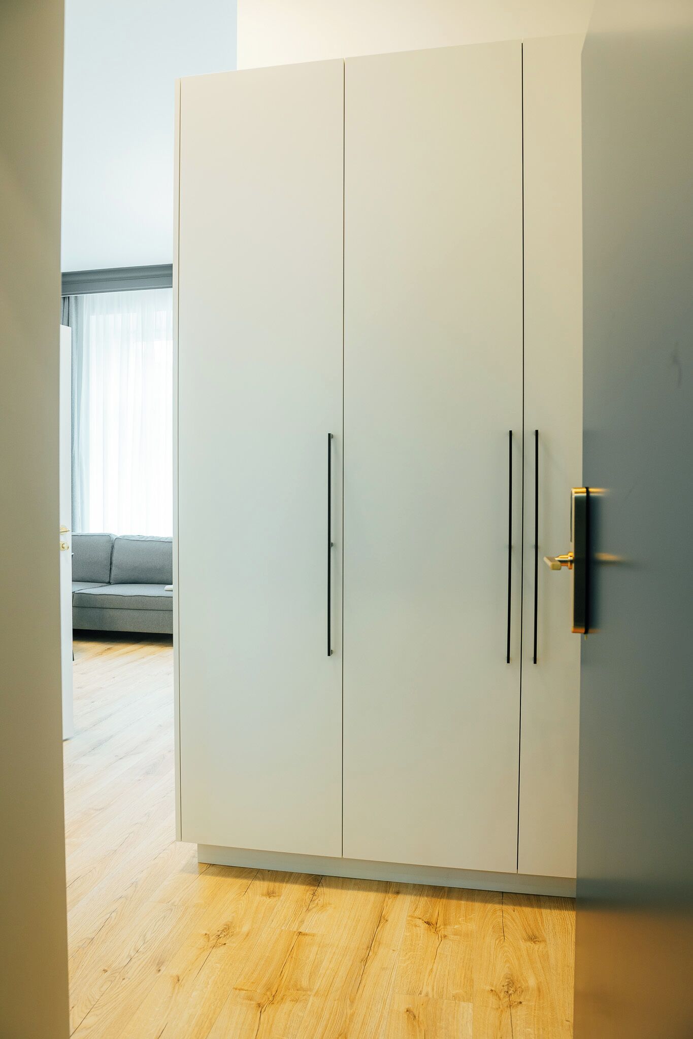 Guarda-roupa moderno com duas portas e puxadores, chão de madeira e um sofá cinzento ao fundo.
