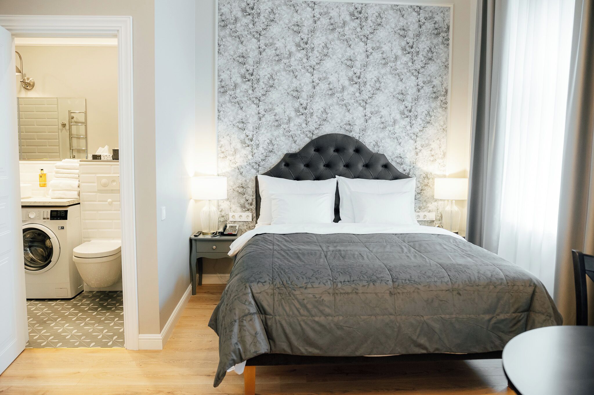 A bedroom with a large bed, a nightstand, a wall-mounted lamp, and a view of a bathroom with a washing machine and toilet.